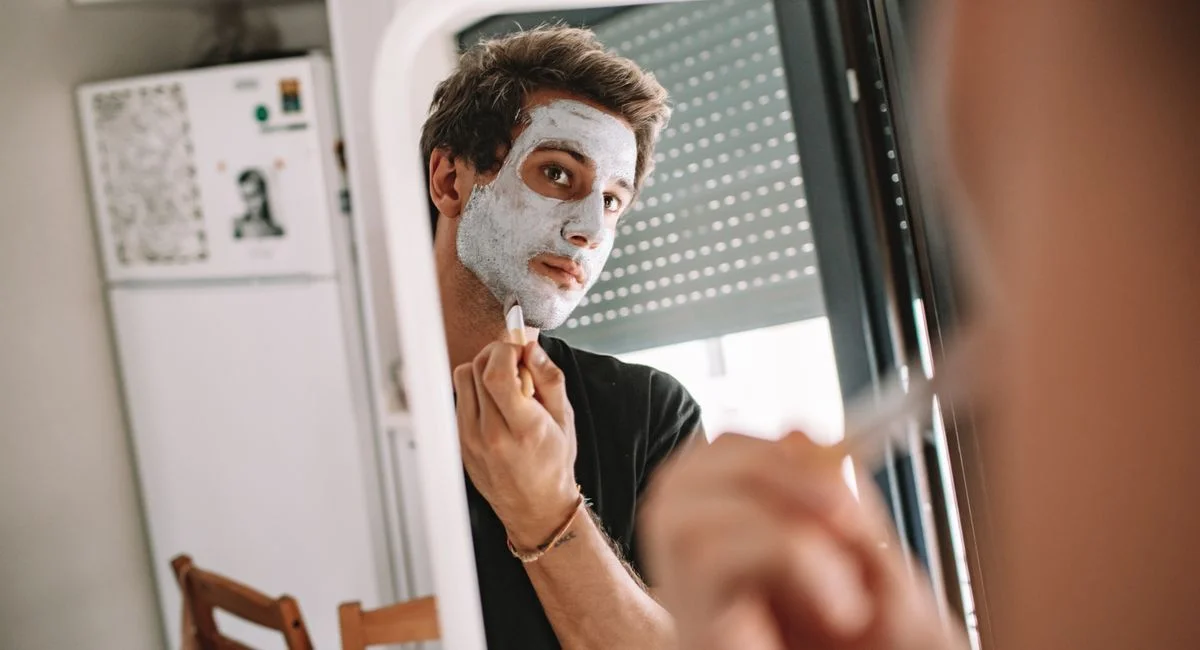 A man applying a face mask while looking into a mirror, emphasizing self-care and grooming.