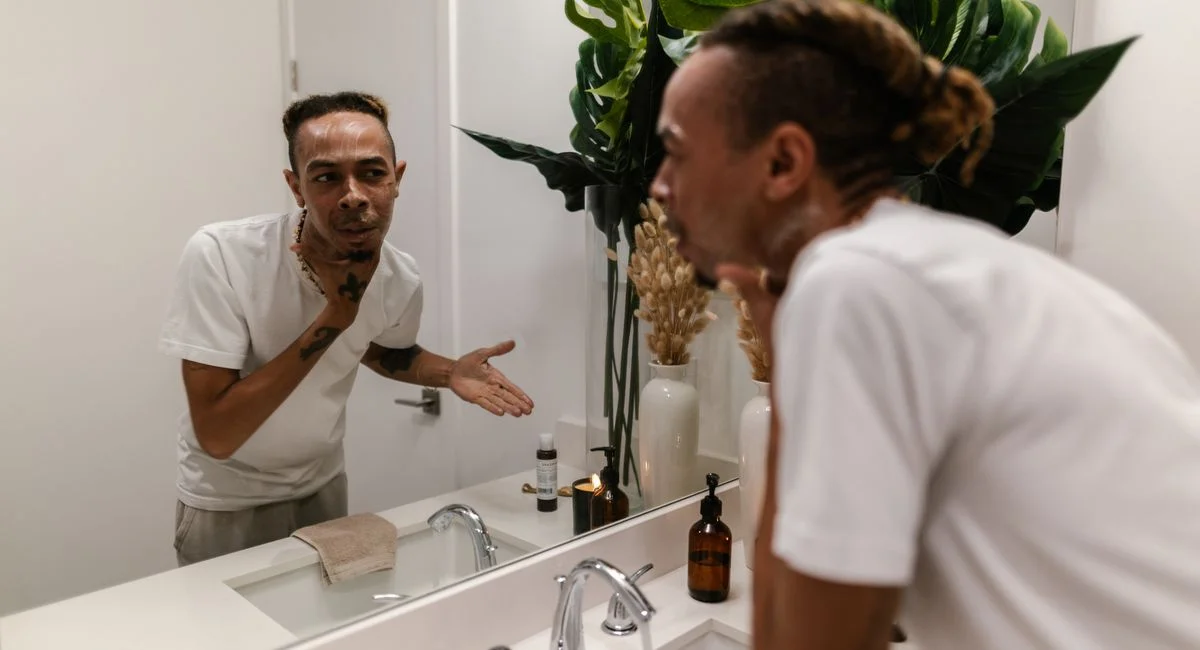 A young man applying skincare product while looking in the mirror in a modern bathroom.