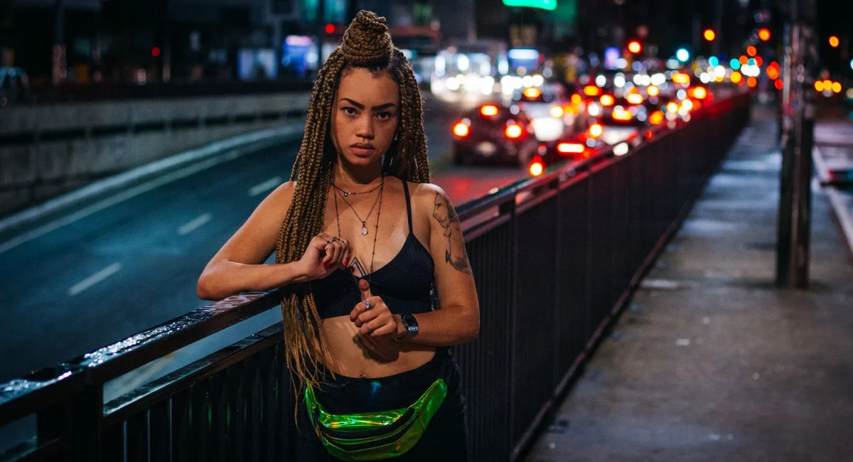 A beautiful woman stands confidently on a city street at night, surrounded by blurred traffic lights.