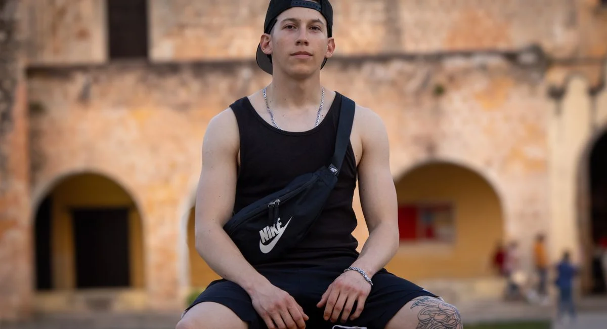 Young man in casual outfit sits in front of a historic building, exuding confidence.