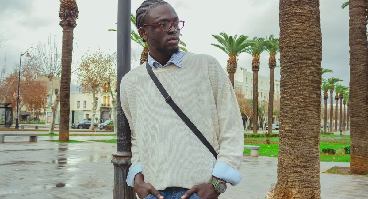 Young man in glasses and casual attire standing near palm trees in a city park.
