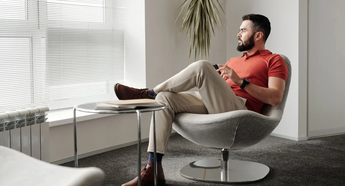 Businessman in a casual setting, looking thoughtful by a window in a modern office chair.