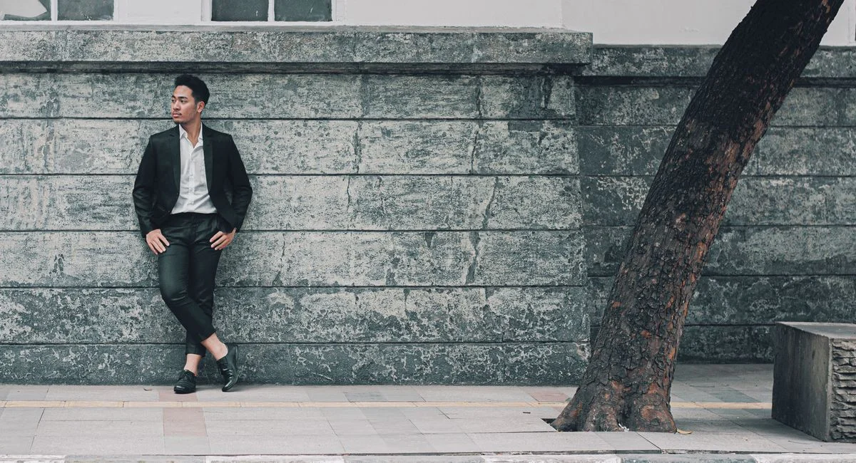 A fashionable man in a blazer leans against a wall in an urban setting, showcasing elegance and style.