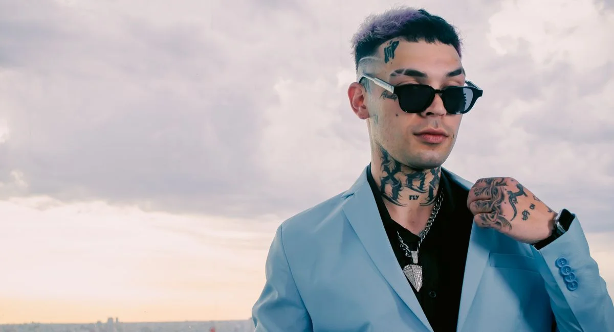 Stylish man in sunglasses and a blue blazer showcasing tattoos, standing outdoors with a cityscape background.