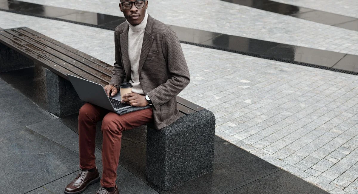 Stylish businessman sitting outdoors on a bench with a laptop and coffee, wearing glasses and a blazer.