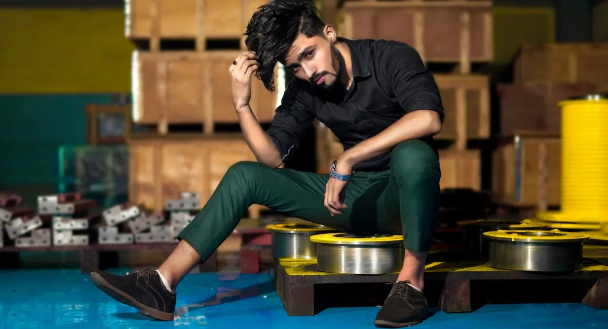 A fashionable man in a black shirt and green pants sits in an industrial setting, exuding modern style.