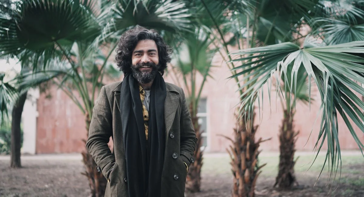A cheerful man with curly hair and a beard stands amidst vibrant palm trees in a garden.