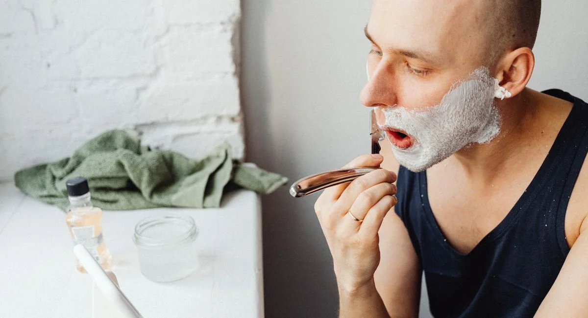 Bald man using a razor and shaving cream in a bathroom setting.