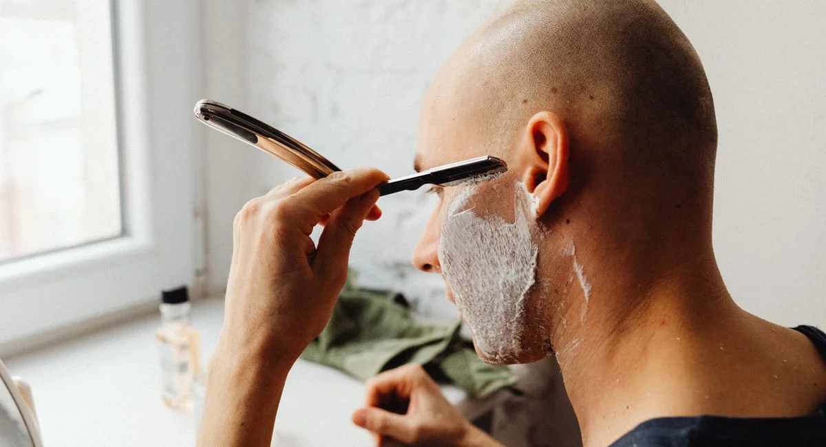 Adult man shaving with straight razor and shaving cream in a light-filled room.