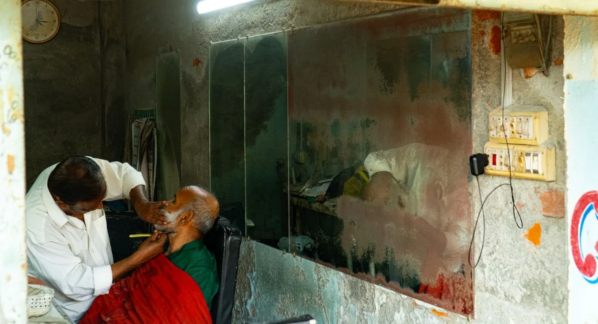 An elderly man receiving a traditional shave at a rustic barber shop seen through a window.
