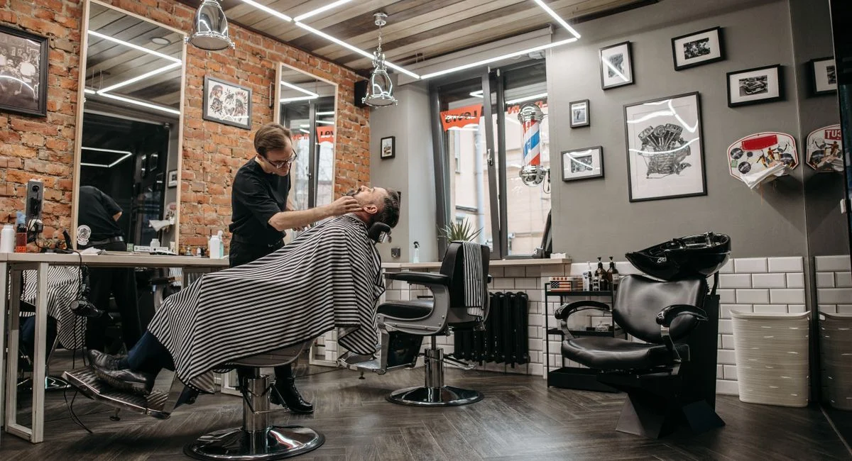 A barber attends to a client in a stylish, contemporary barbershop with brick walls and framed art.