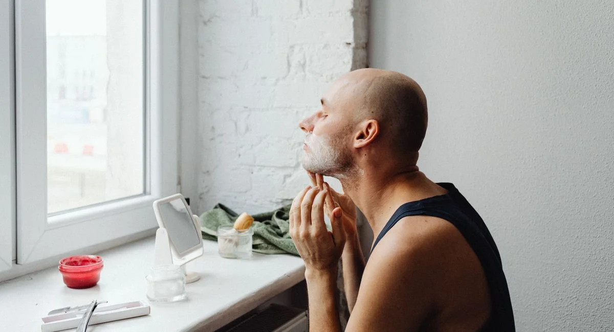 A bald man applying shaving cream to his chin while looking out the window