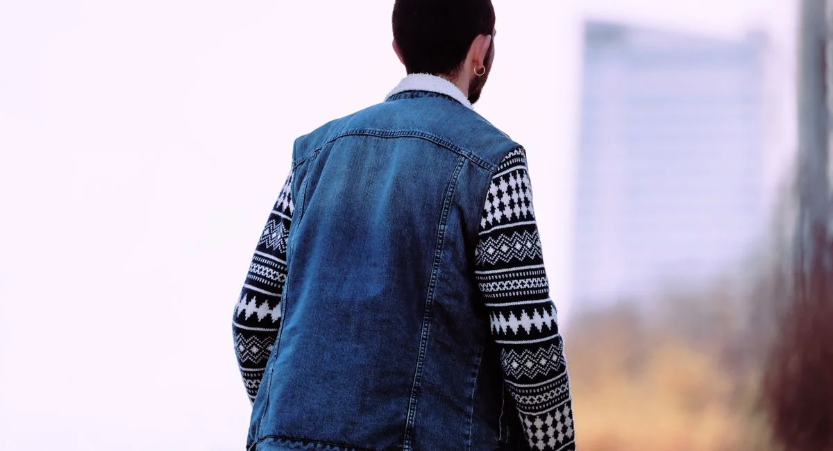 A man in a denim vest and patterned sweater gazes at a blurred cityscape in the background.