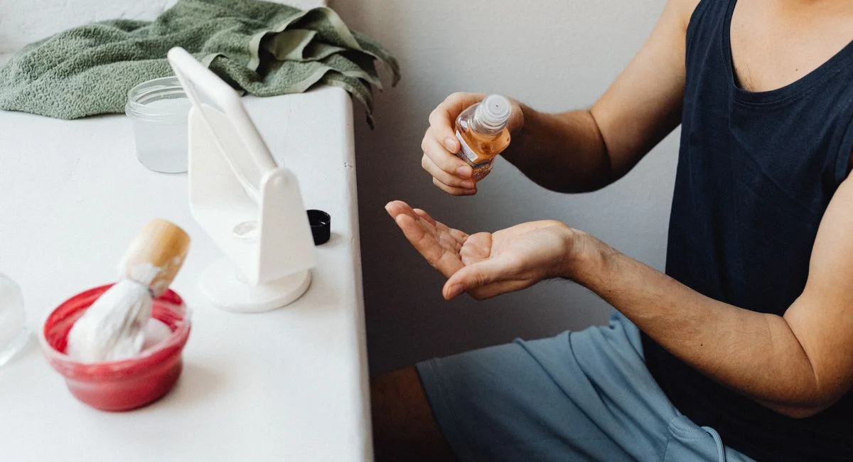 Adult man applying aftershave in a casual bathroom setup with mirror and shaving accessories.