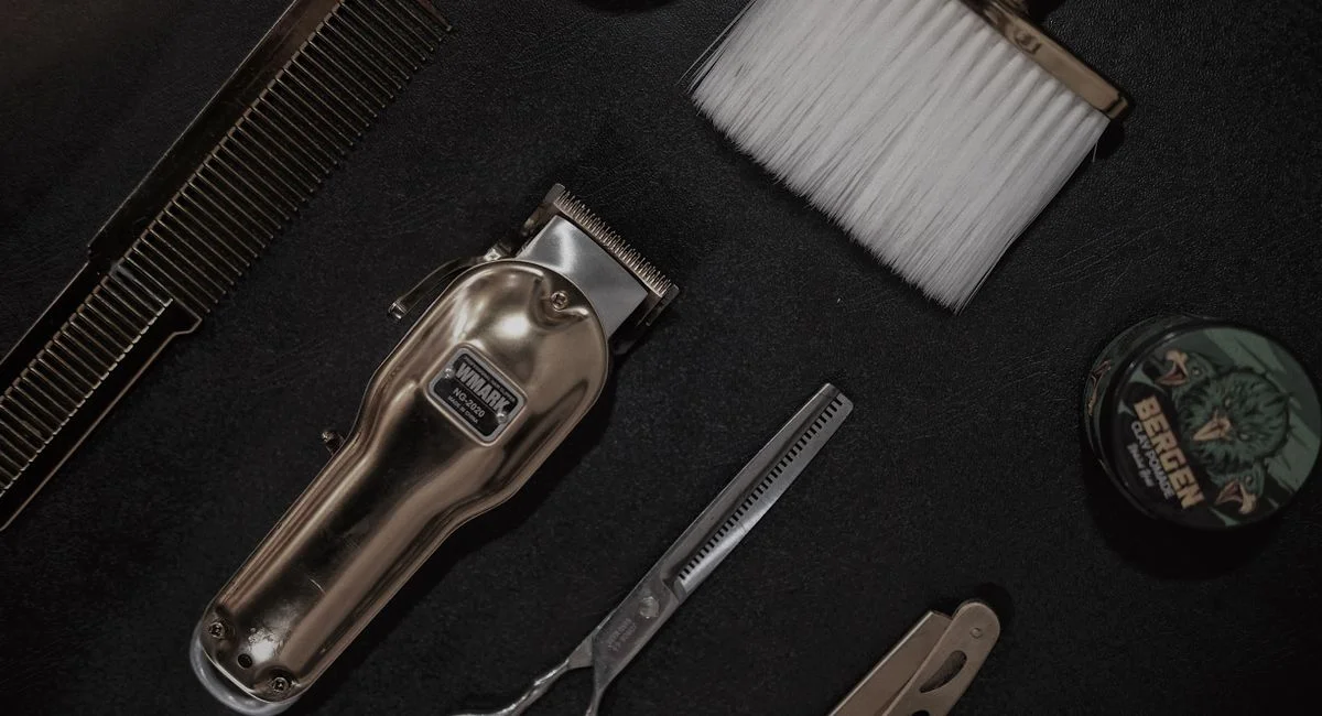 Flat lay of barber tools including clipper, scissors, and comb on a black surface.