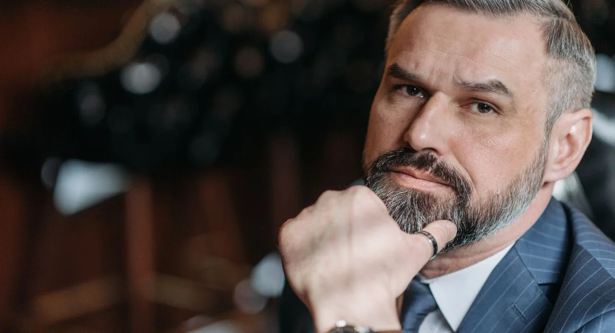 A thoughtful, bearded man in a suit poses indoors, exuding style and confidence.