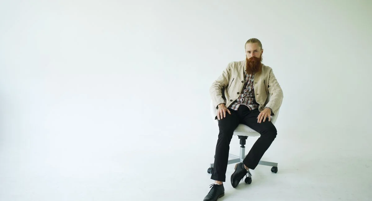 Bearded man in casual blazer and plaid shirt seated on a chair in a bright, modern setting.