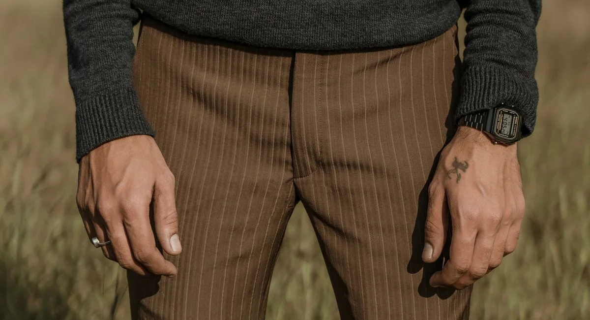 Man's hands with a tattoo and wristwatch wearing brown pinstripe trousers in an outdoor setting.