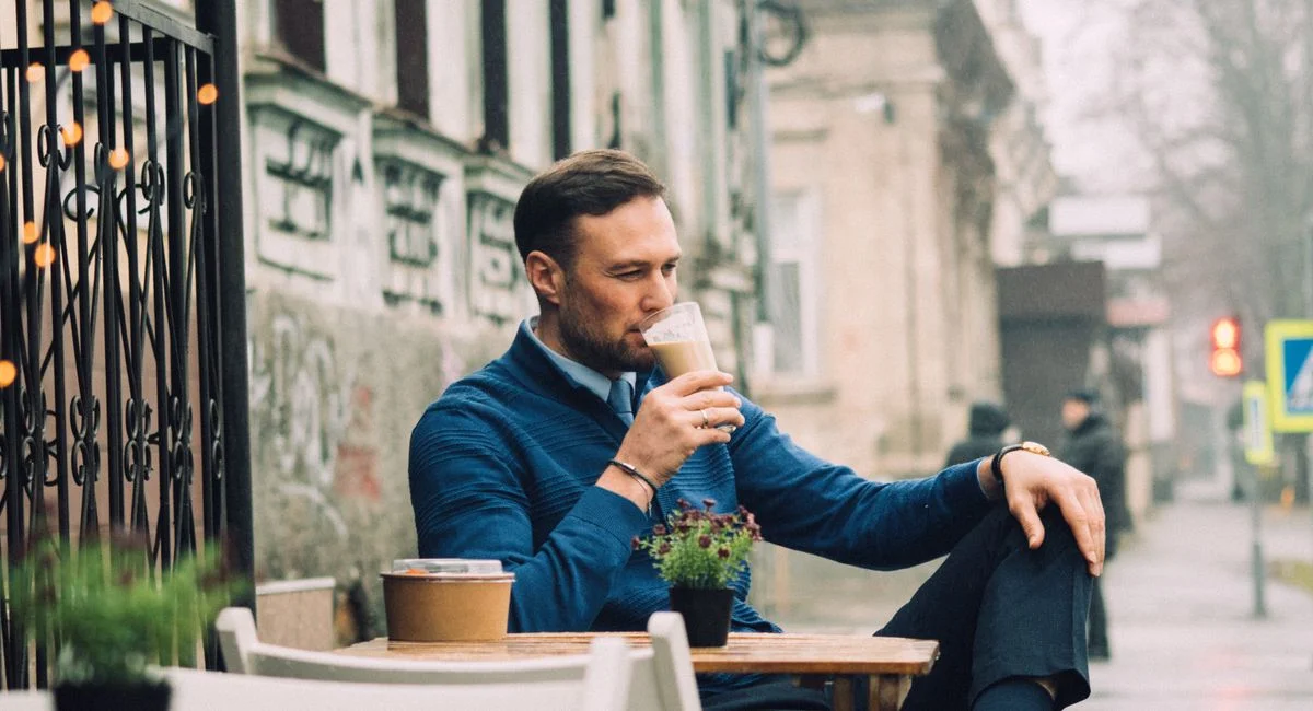 Young man sipping coffee at an outdoor cafe, enjoying a relaxing moment in a lively urban setting.