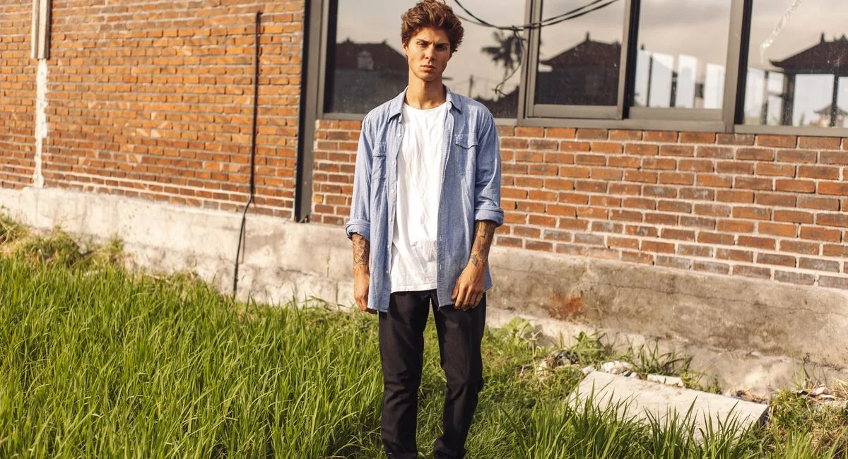 Young man in casual attire stands outdoors by a brick building, showcasing tattoos.