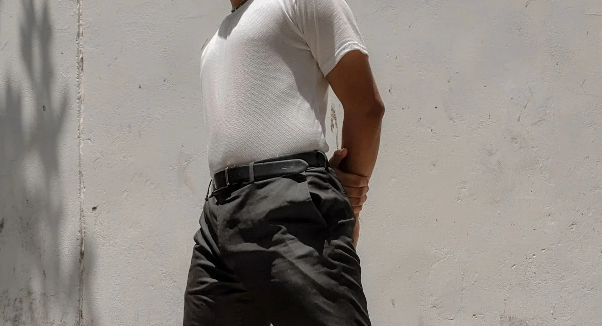 Stylish man in white t-shirt and dark pants posing confidently outdoors against a textured wall.