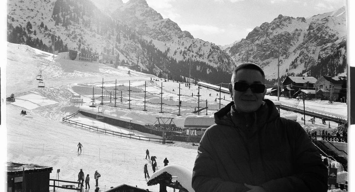 Black and white photo of a ski resort with mountains, people skiing, and a man in sunglasses.