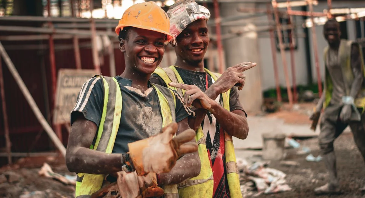 Two smiling construction workers in hard hats and safety vests pointing with joy on a construction site.