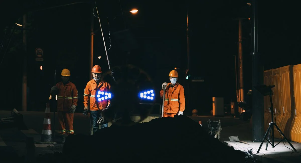 Workers in reflective gear and helmets conduct roadworks at night under bright lights.
