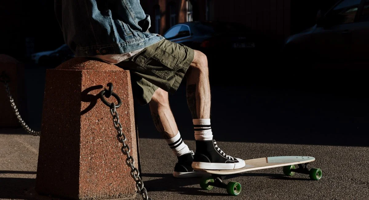 A person sits on a skateboard in an urban setting, dressed in casual attire.