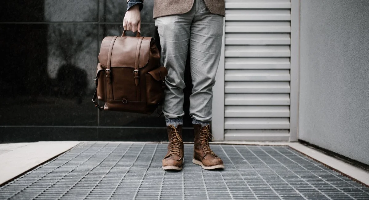 A man in stylish attire holding a leather backpack outdoors.