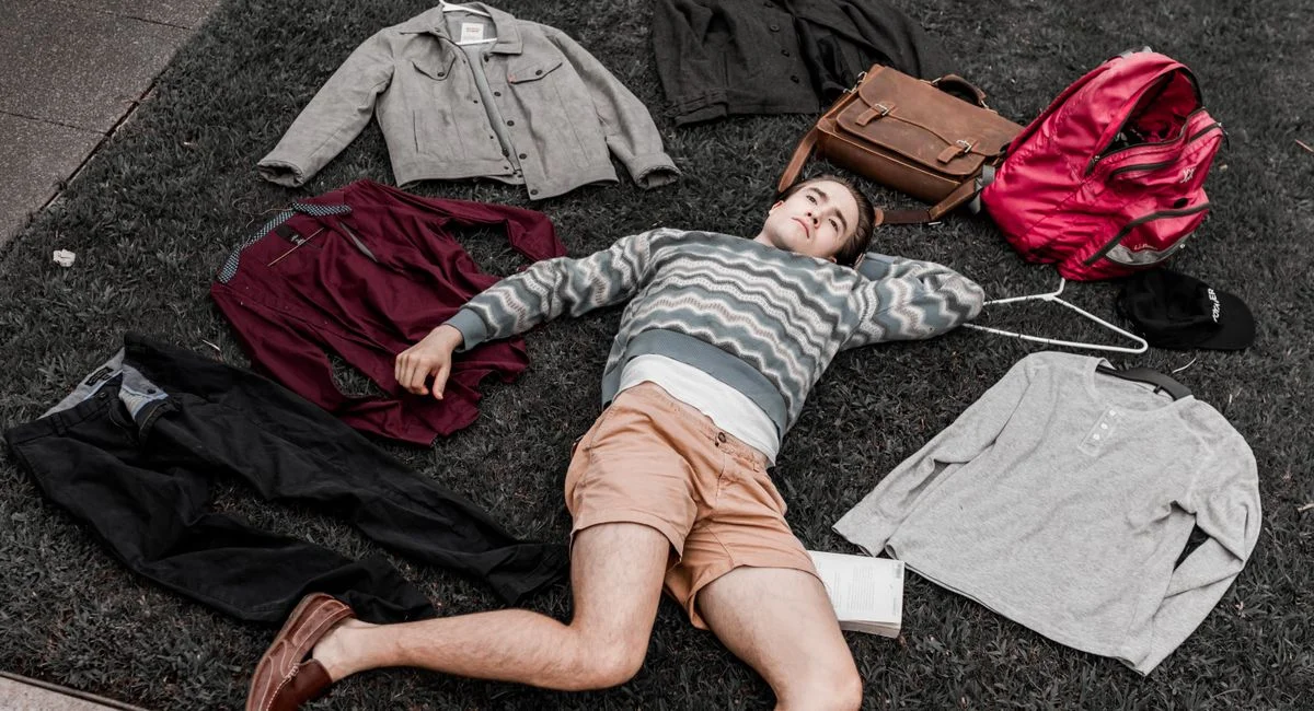 A young man lies on grass surrounded by clothes and bags in an outdoor setting.