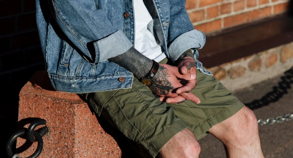 Close-up of a tattooed man sitting on a bollard. Casual denim and shorts style.