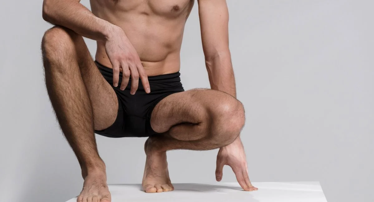 Close-up of a shirtless man squatting, wearing black underwear on a white platform.