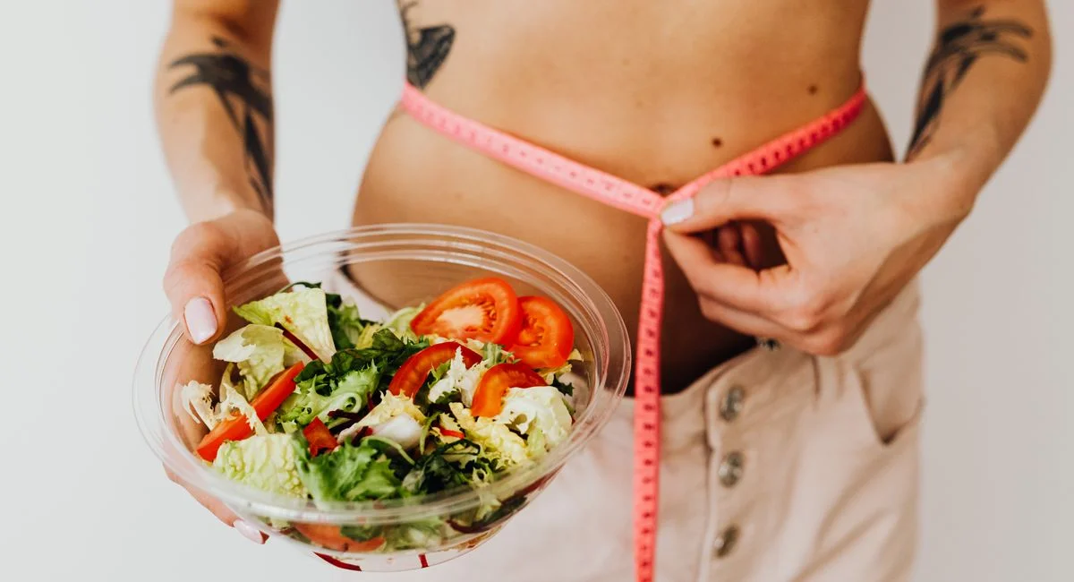 Woman holding vegetable salad and measuring waist with tape, symbolizing healthy lifestyle choices.