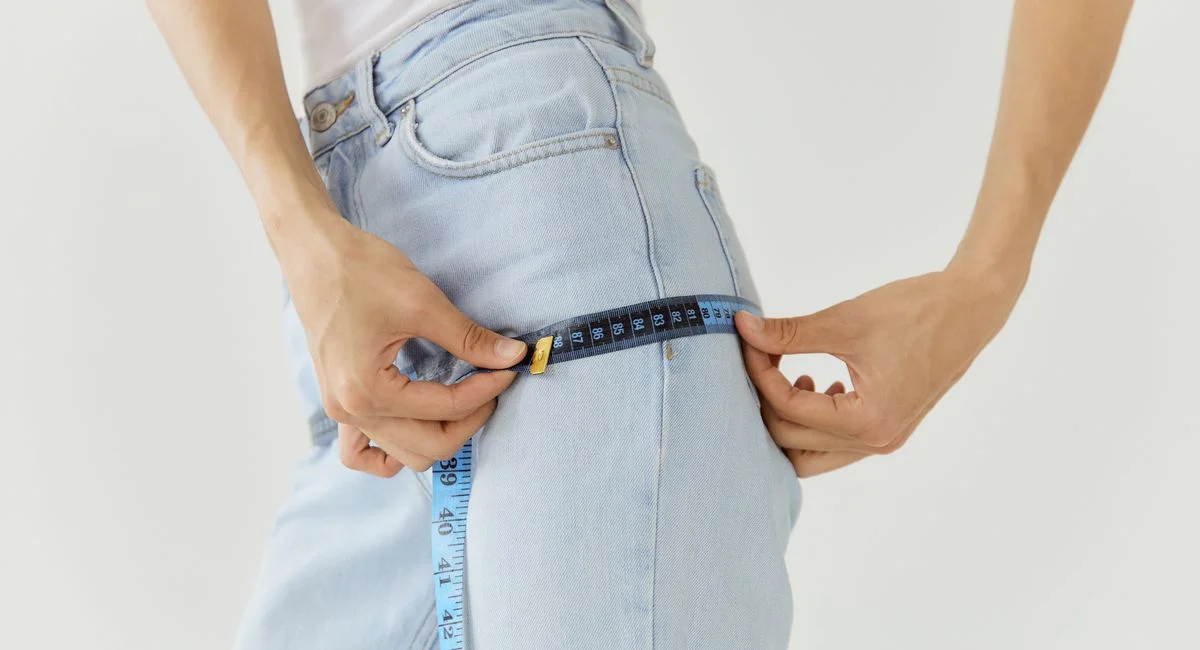 Person measuring denim pants waist with a blue tape measure on a white background.