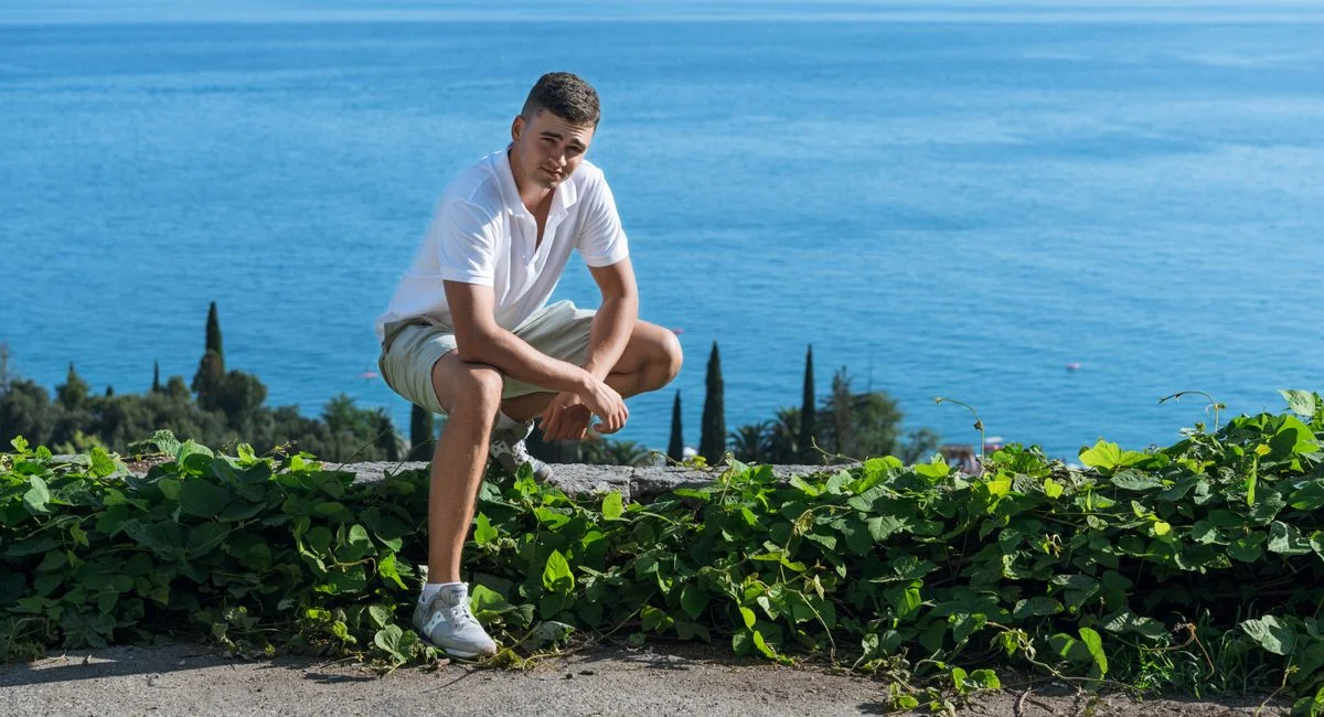 A young man crouching by the ocean, surrounded by greenery, enjoying a sunny day outdoors.
