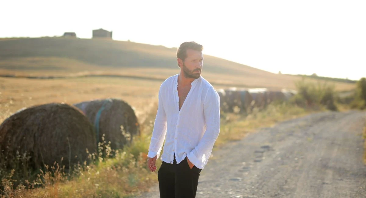 A man in a white shirt walks along a rural dirt road beside hay bales during sunset.