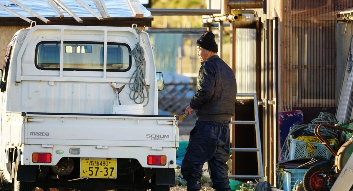 Elderly fisherman working by his truck with ropes and crates outdoors in Japan.