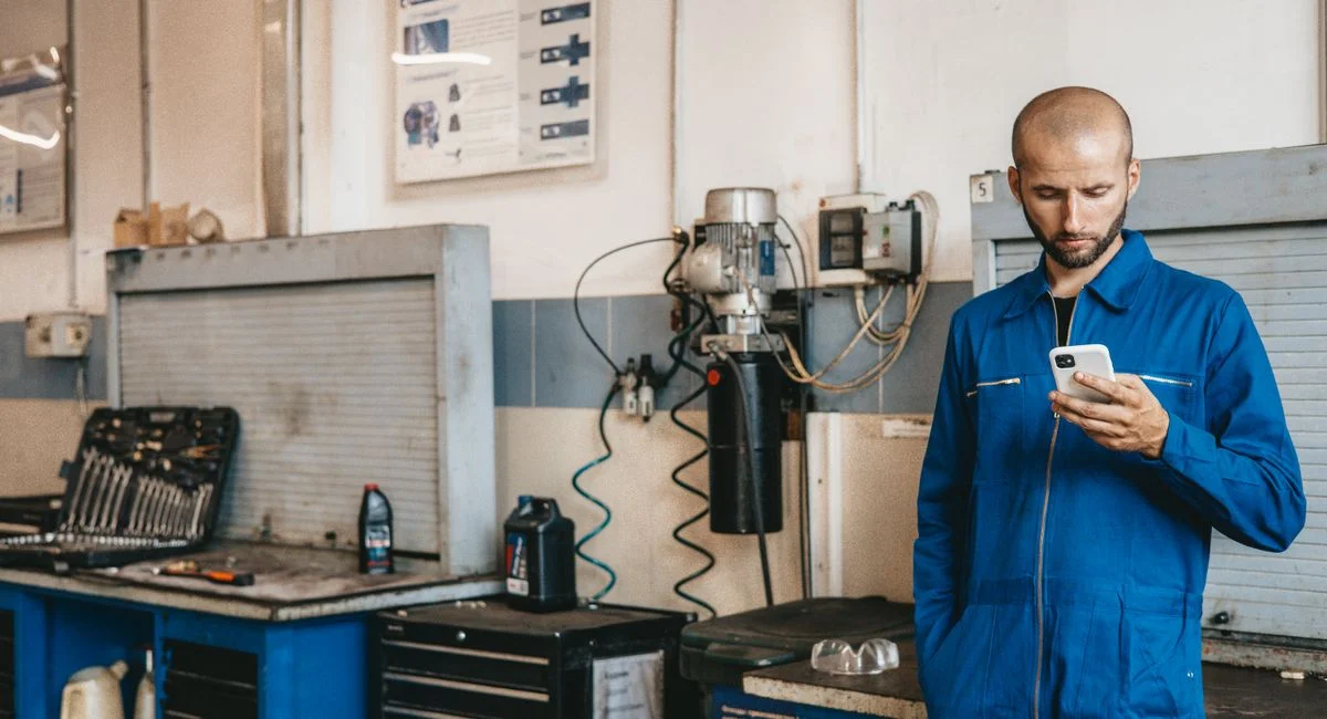 Mechanic in blue coveralls using a smartphone in an auto repair workshop.